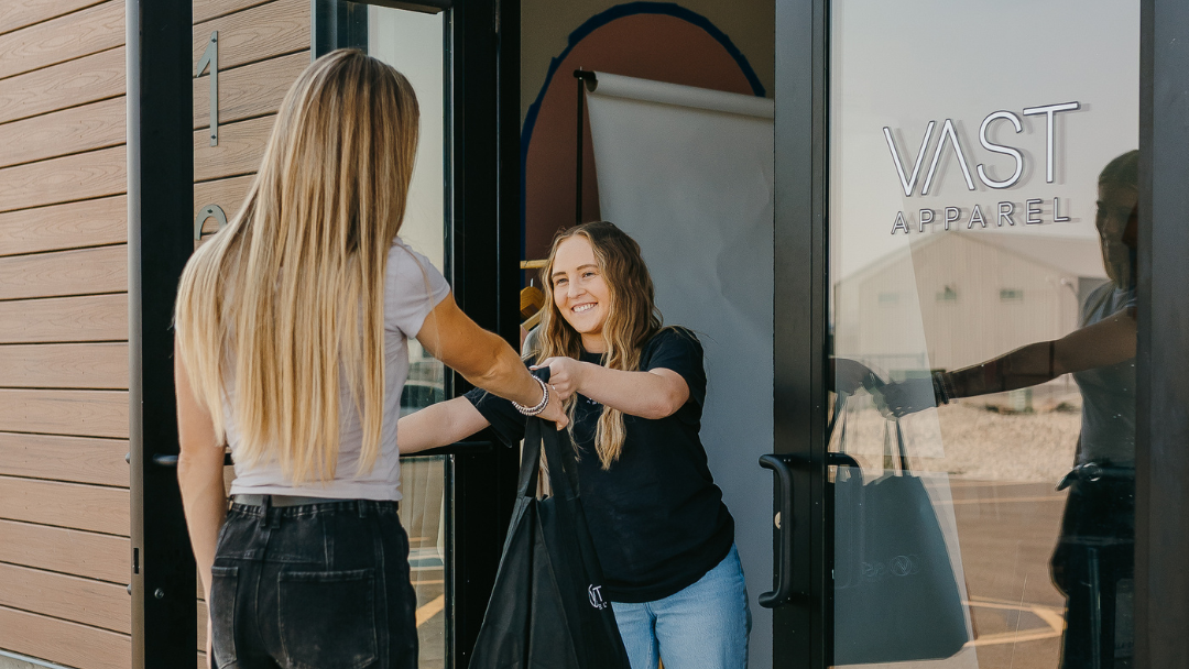Two women shaking hands outside a store with 'Vast Apparel' branding.